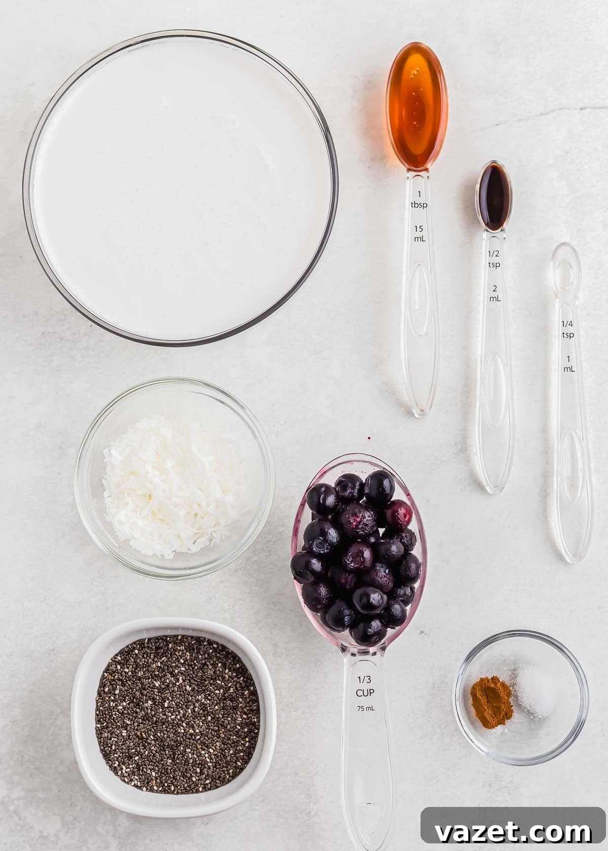 Berry Burst Chia Delight 4 Clear bowls and measuring spoons filled with various ingredients including coconut milk, honey, vanilla extract, shredded coconut, blueberries, chia seeds, salt, and cinnamon laid out on a white surface.