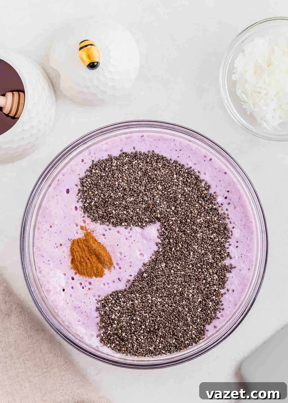 Berry Burst Chia Delight 5 A glass bowl filled with a blended blueberry mixture topped with a large amount of chia seeds arranged in a swirling shape, a small pile of cinnamon, with honey and shredded coconut nearby.