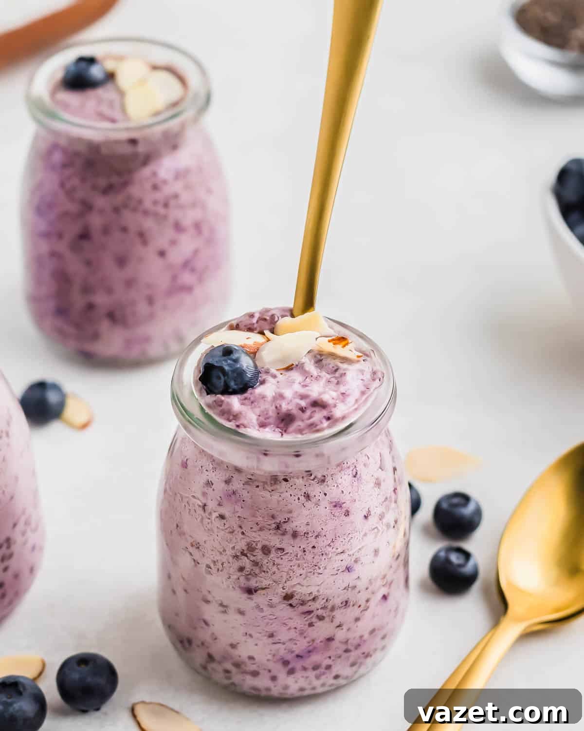 Berry Burst Chia Delight 8 Three jars of blueberry chia pudding topped with fresh blueberries and almond slices, with a golden spoon inserted in one. Scattered blueberries and almond slices are on the surface.