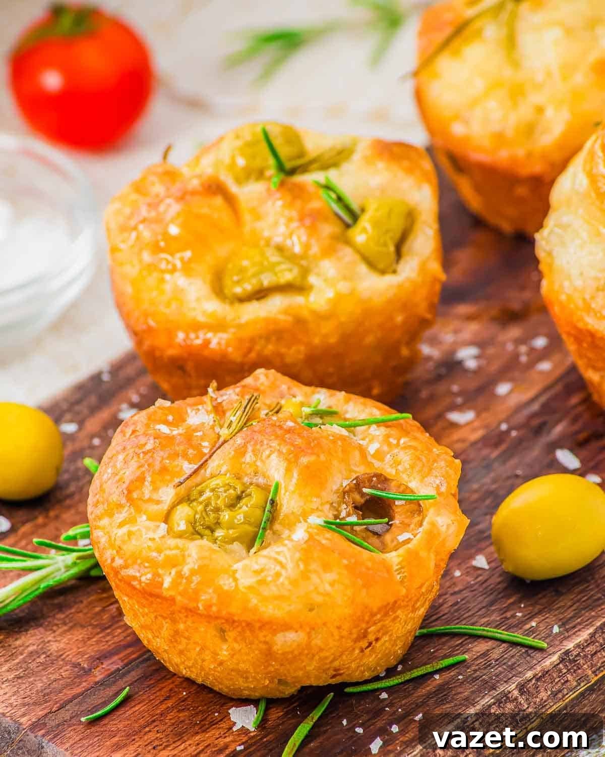 Golden baked focaccia muffins topped with green olives and rosemary on a wooden board.