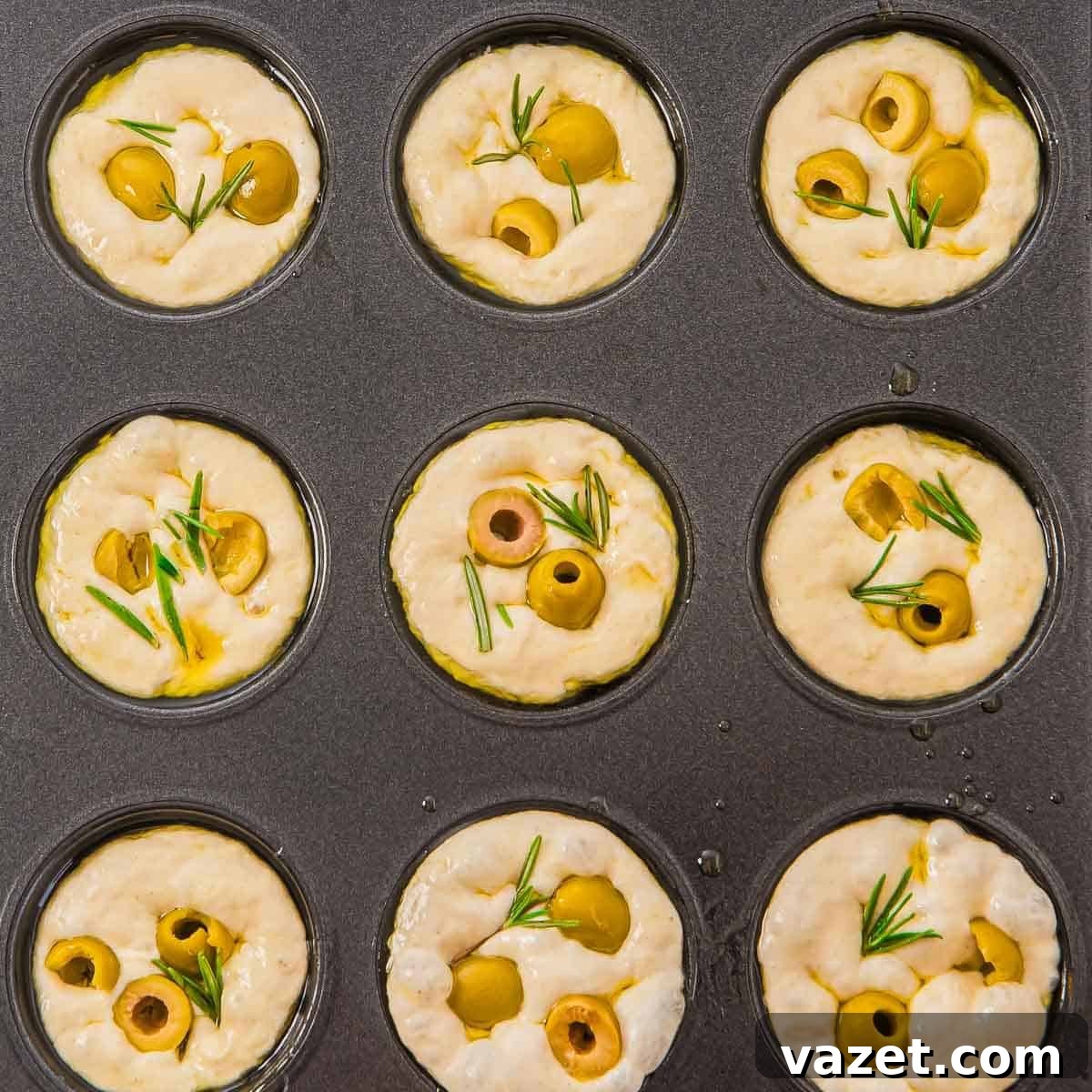 Muffin tin filled with dough topped with olives and rosemary, ready to bake.