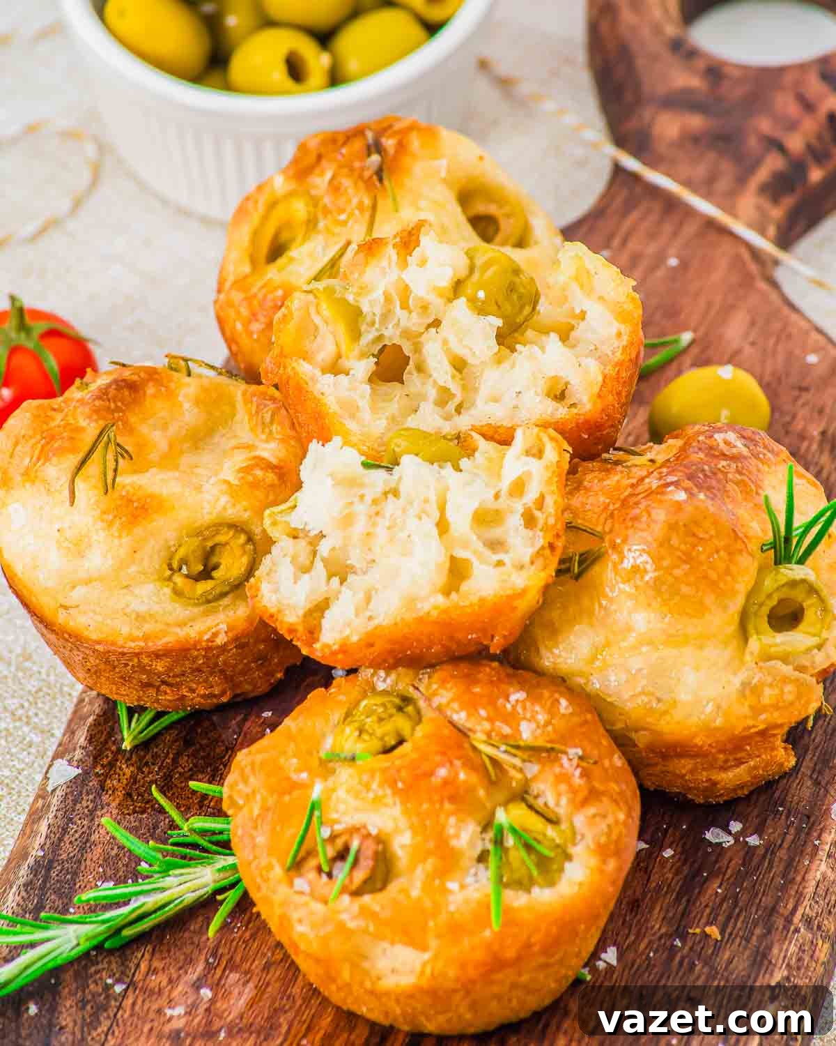 Focaccia muffins stacked on a wooden board with olives and rosemary.