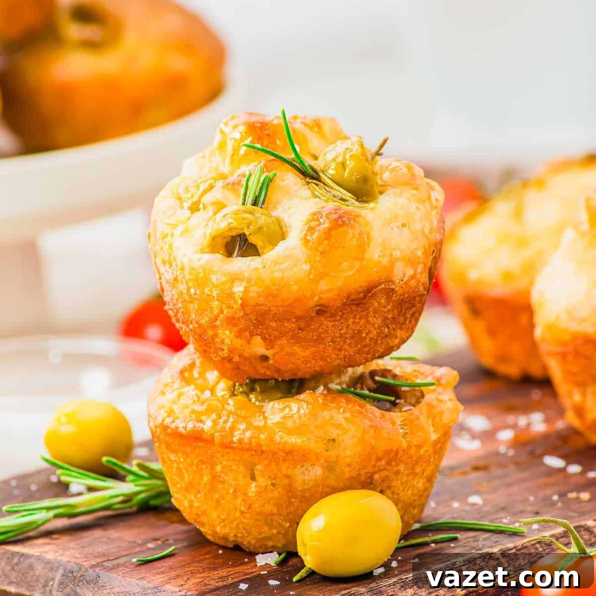 Two focaccia muffins topped with rosemary and green olives stacked on a wooden board.