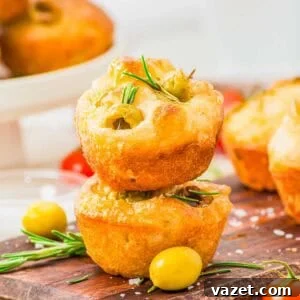Two focaccia muffins topped with rosemary and green olives are stacked on a wooden board