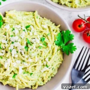 A bowl of creamy green pasta garnished with crumbled white cheese and fresh parsley, with a sprig of parsley and cluster of cherry tomatoes nearby. Black forks are placed beside the bowl on a white surface.