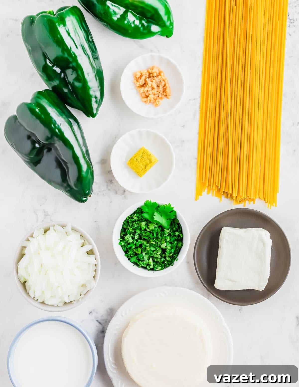 All the fresh ingredients needed to make Espagueti Verde laid out on a white surface, including poblano peppers, uncooked spaghetti, onion, cilantro, garlic, bouillon cube, cream cheese, Mexican crema, and queso fresco.