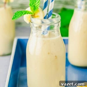 Korean Banana Bliss 10 A glass bottle filled with creamy milk, garnished with a banana slice and mint sprig. Two striped straws are in the bottle, which is placed on a blue tray with another smoothie in the background.