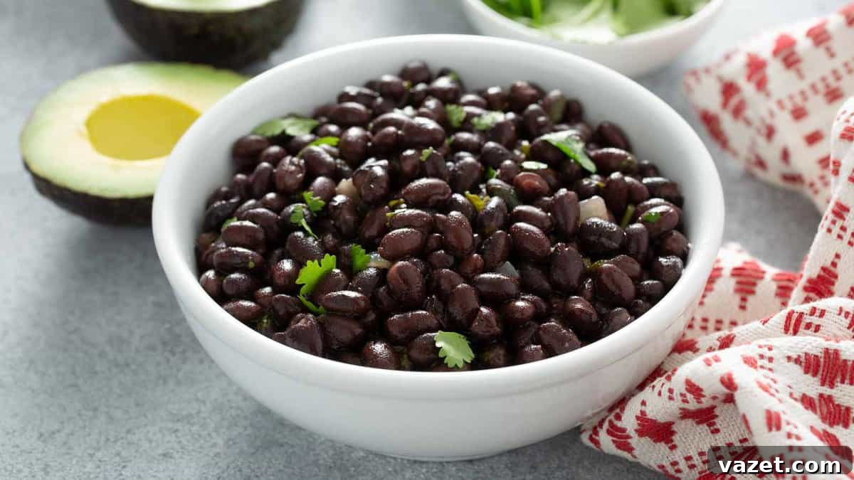A white bowl filled with perfectly cooked black beans garnished with fresh cilantro, positioned on a sleek grey surface next to half an avocado and a rustic red-and-white striped cloth. The composition highlights the vibrant colors and fresh ingredients of a healthy meal.