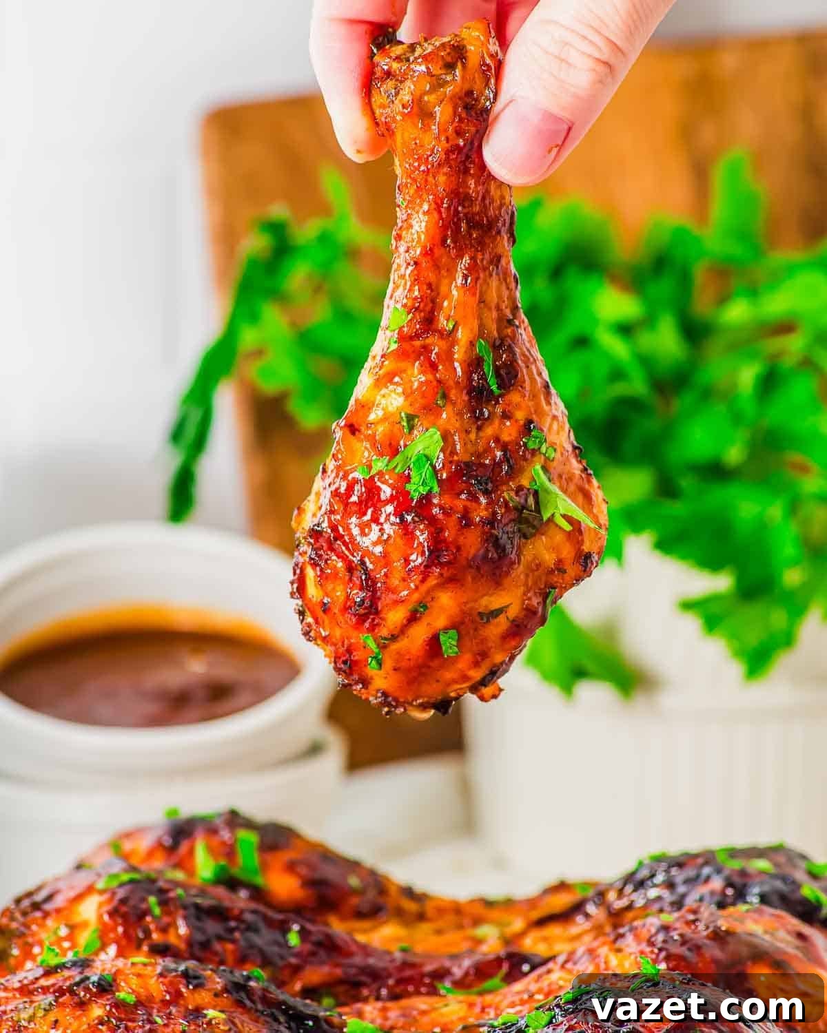 A hand holds up a glazed, crispy air fried chicken drumstick garnished with chopped herbs. In the background, there is a bowl of dipping sauce and a bunch of fresh parsley.