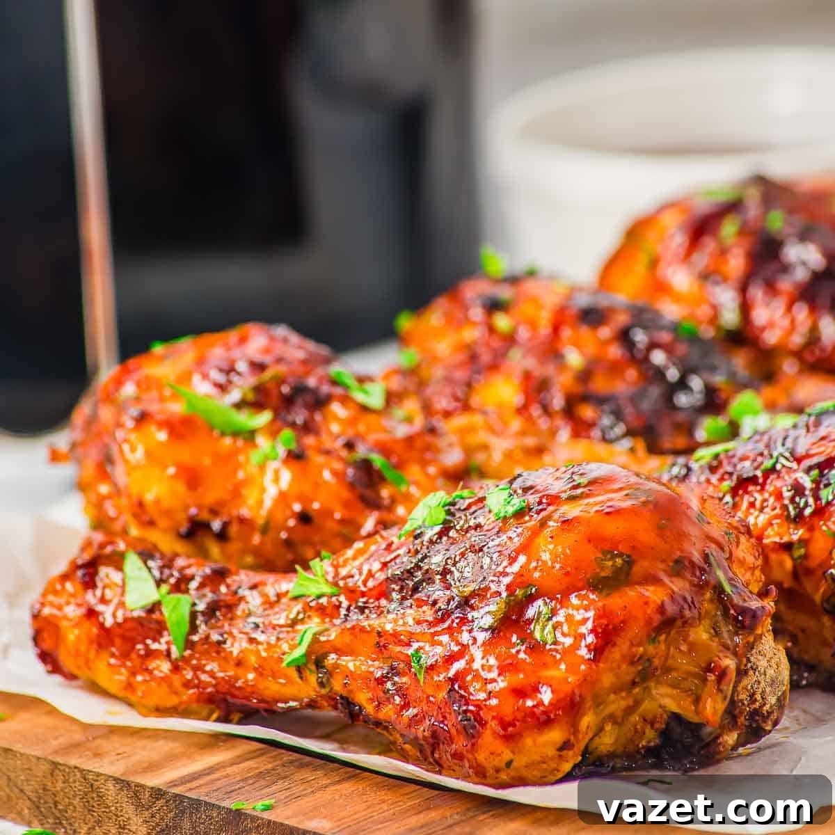 Close-up of BBQ glazed, air fryer chicken drumsticks garnished with chopped fresh herbs, arranged on a wooden board lined with parchment paper. The chicken has a shiny, caramelized surface.
