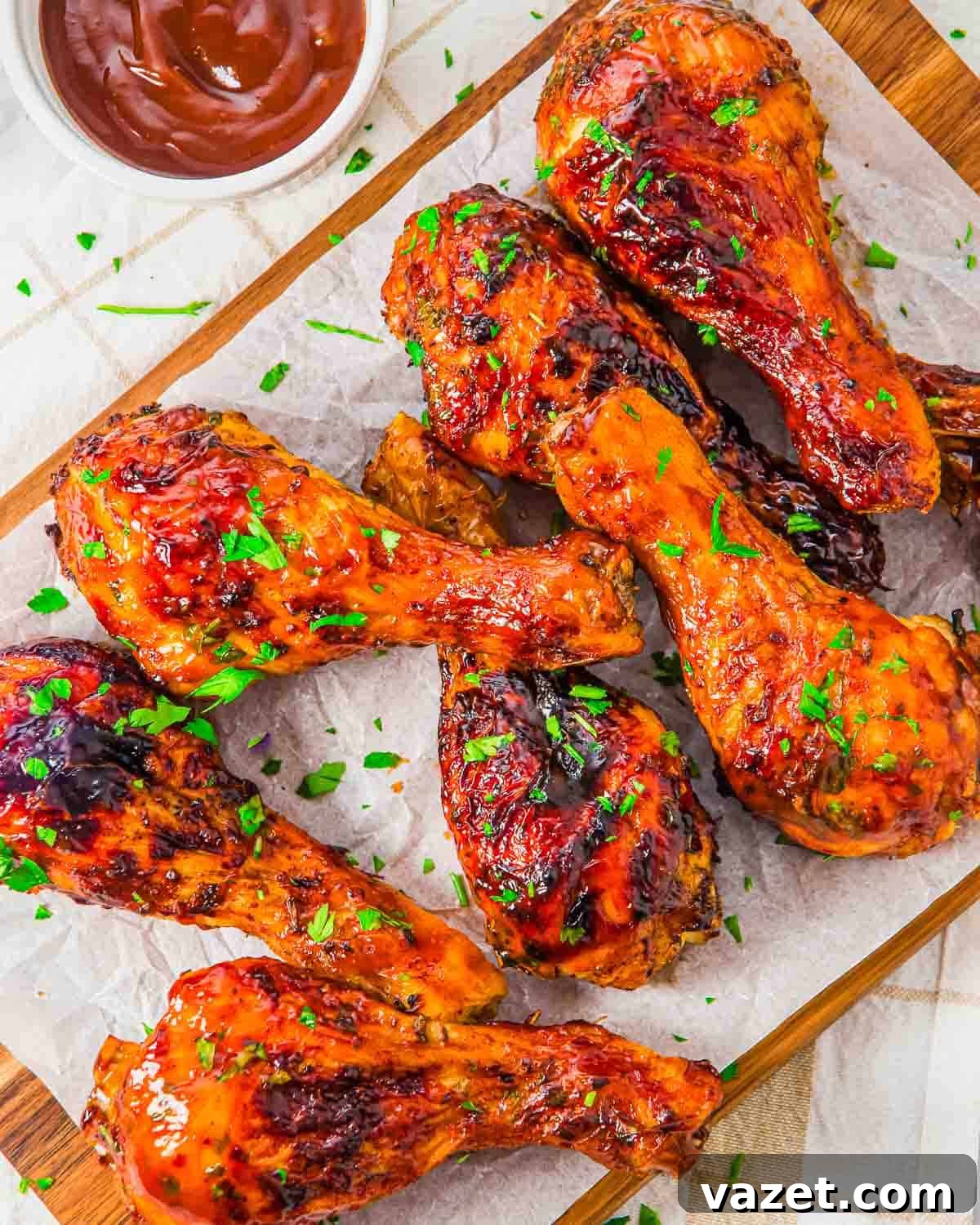 Air fried chicken drumsticks glazed with barbecue sauce, garnished with chopped parsley, arranged on parchment paper atop a wooden board, with a small bowl of barbecue sauce nearby.