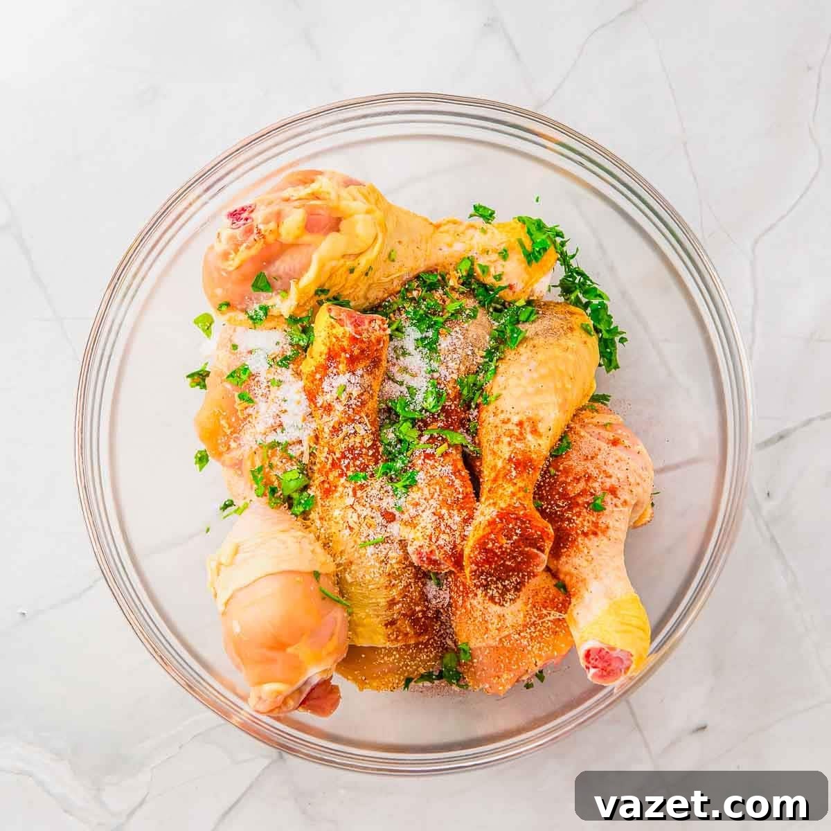 Raw chicken drumsticks in a glass bowl, sprinkled with chopped fresh herbs, salt, and seasoning, set on a light-colored marble surface.