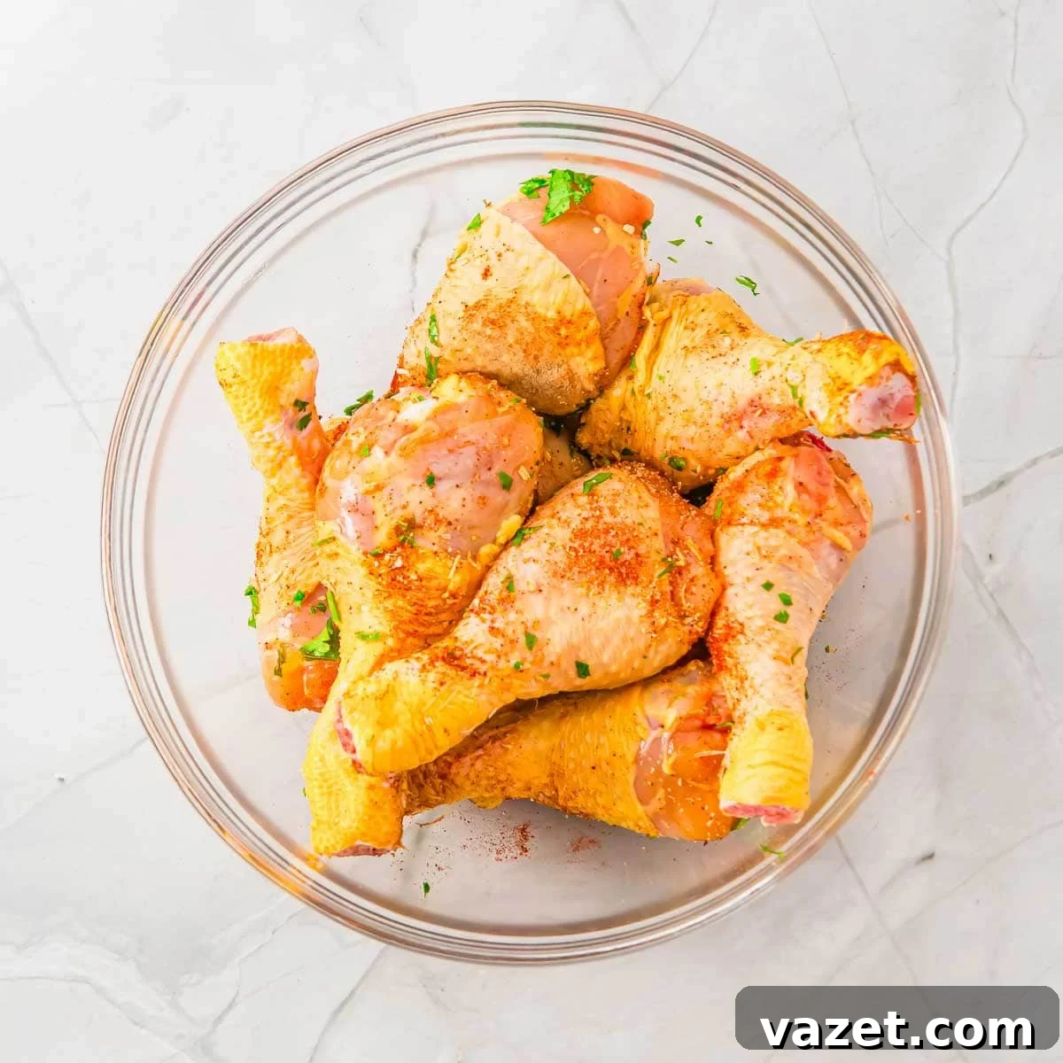 A glass bowl filled with raw chicken drumsticks, sprinkled with seasoning and chopped herbs, sits on a light-colored marble surface.
