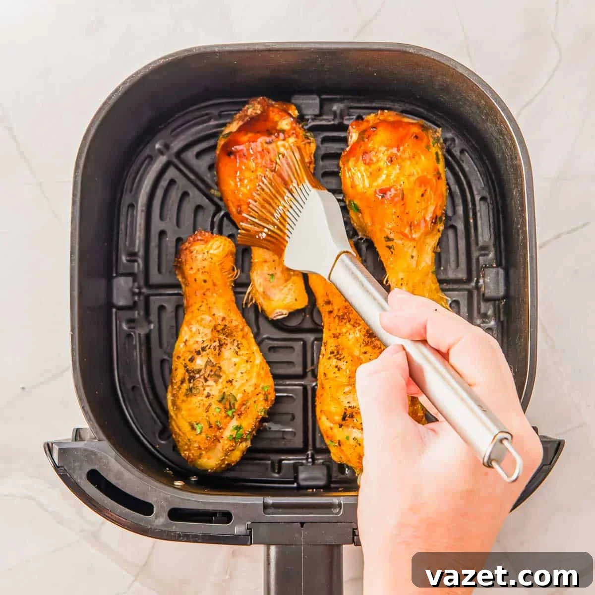 A hand uses a brush to apply BBQ sauce to cooked chicken drumsticks inside an air fryer basket on a light-colored surface.