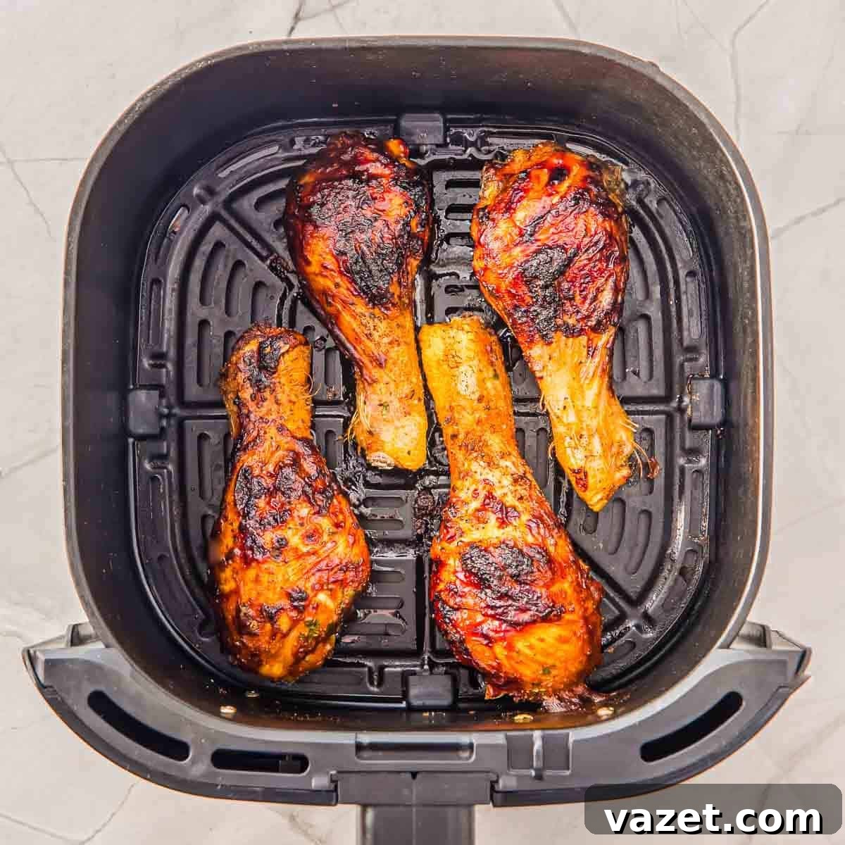 Four cooked, slightly charred chicken drumsticks in an air fryer basket on a light marble countertop.