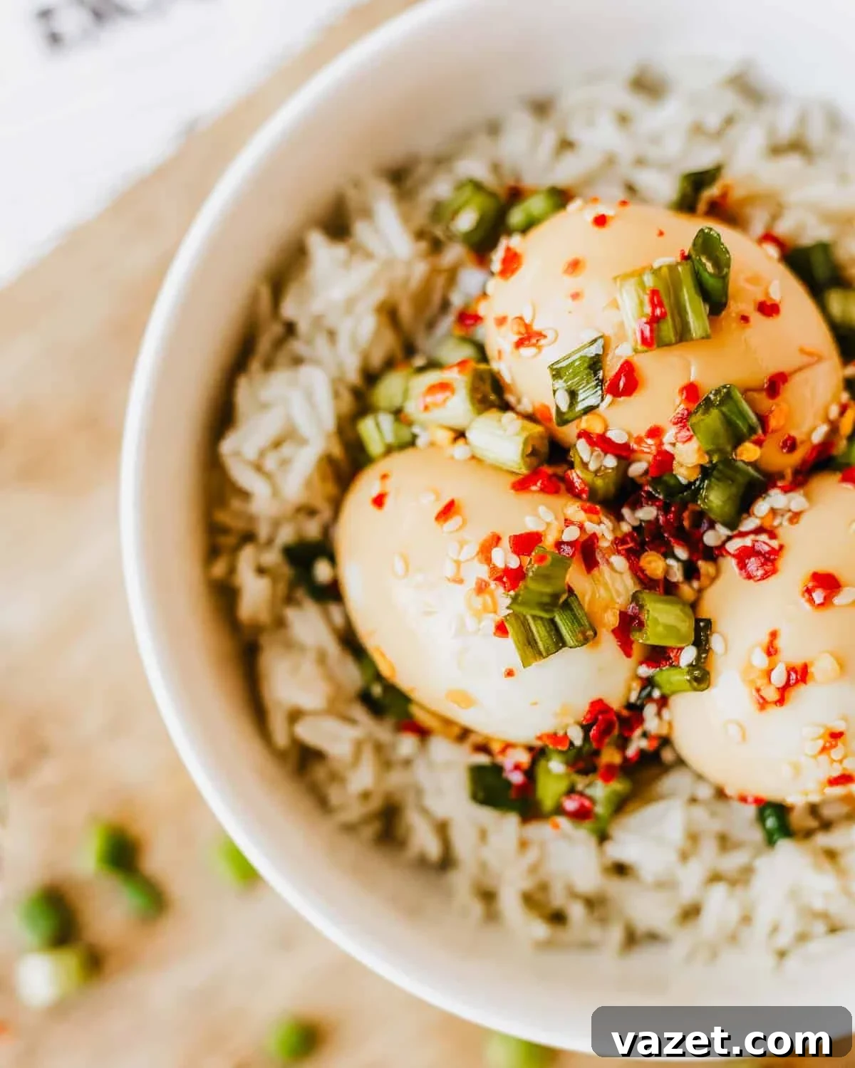 A bowl of white rice topped with three soy sauce marinated eggs, garnished with chopped green onions, red chili flakes, and sesame seeds.