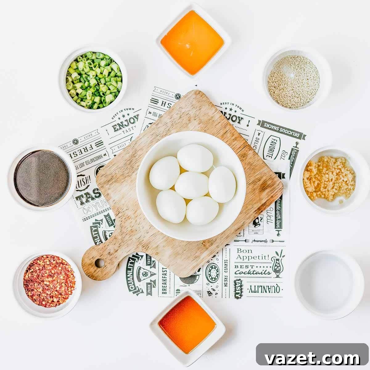 Overhead view of a cutting board with six boiled eggs in a bowl, surrounded by small bowls of chopped green onions, sesame seeds, minced garlic, black pepper, red pepper flakes, and two orange sauces on a white background.