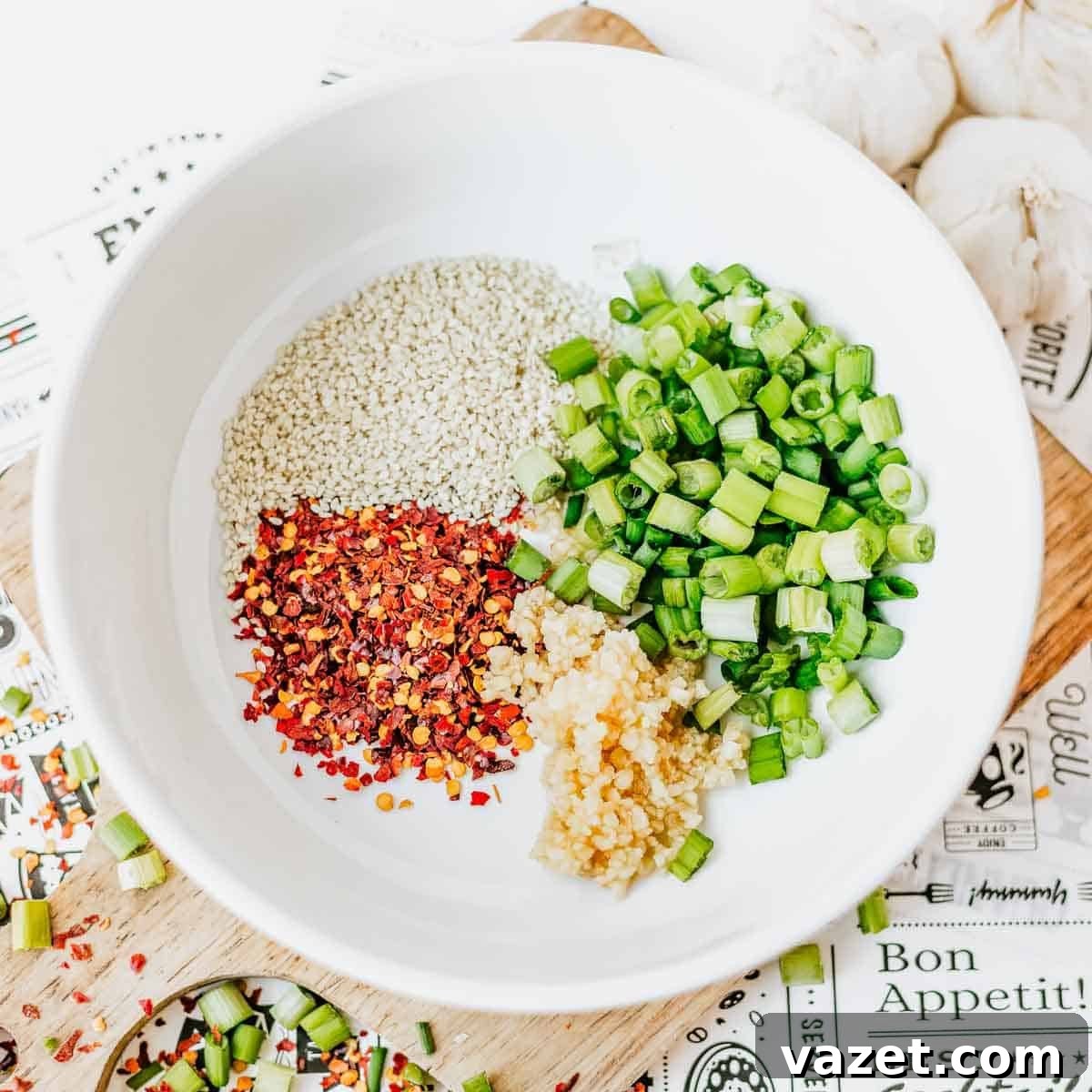 A white bowl contains sesame seeds, red pepper flakes, chopped green onions, and minced garlic, arranged in separate sections. The bowl sits on a wooden surface with scattered green onion slices and a garlic bulb nearby.
