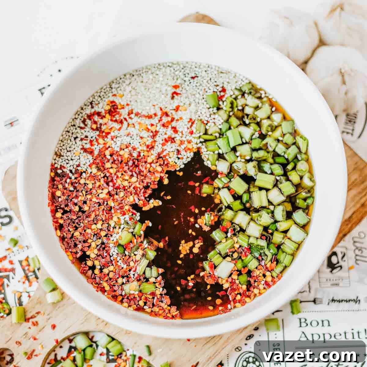 A white bowl filled with a soy sauce mixture containing red chili flakes, sesame seeds, chopped green onions, and a dark liquid, placed on a patterned surface with garlic cloves nearby.