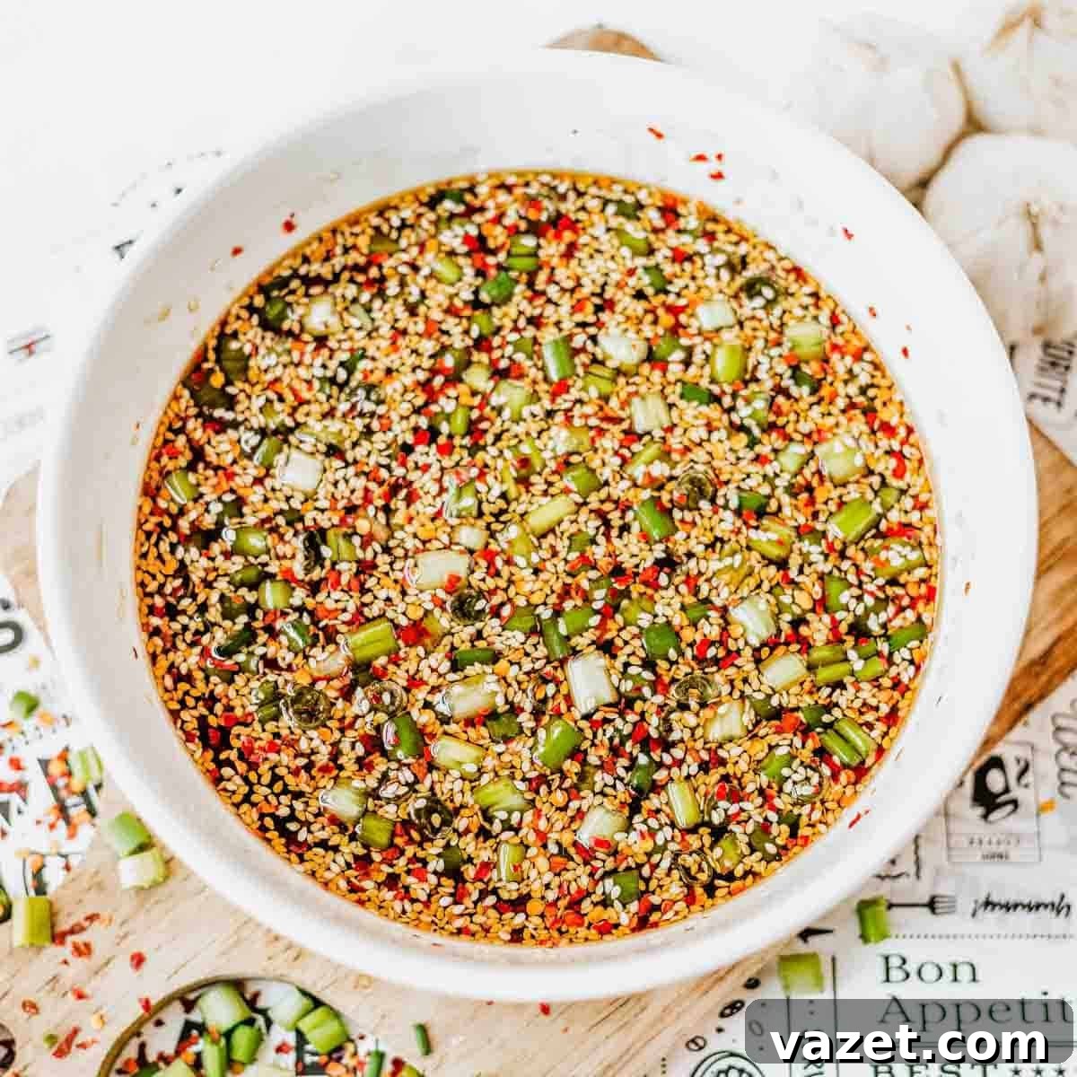 A white bowl filled with a colorful soy sauce marinade containing chopped green onions, sesame seeds, red pepper flakes, and other seasonings. Nearby are pieces of garlic and a cutting board with scattered onions and seasonings.