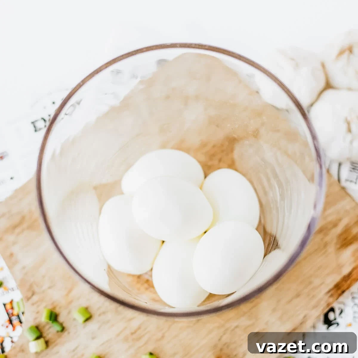 A glass bowl containing five peeled hard-boiled eggs sits on a wooden cutting board next to chopped green onions and garlic cloves.
