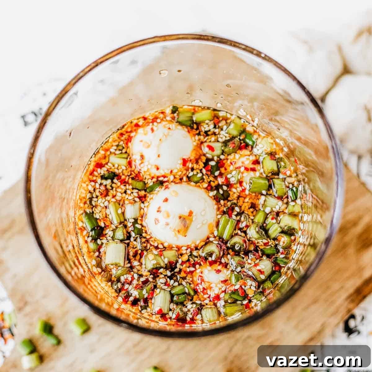 A glass bowl filled with marinated boiled eggs, chopped green onions, sesame seeds, and red chili flakes in a soy-based sauce, placed on a wooden surface.