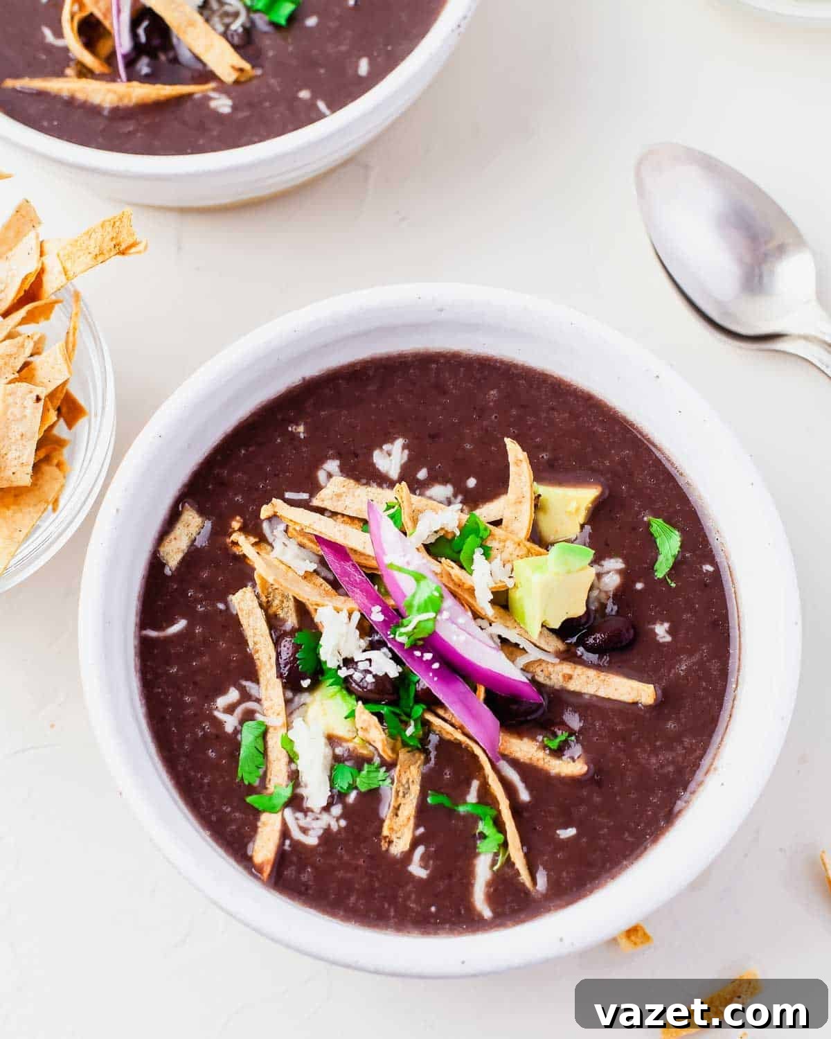 A vibrant bowl of Instant Pot Black Bean Soup generously topped with crispy tortilla strips, fresh avocado slices, finely diced red onion, grated cheese, vibrant cilantro, and a few whole black beans for texture. A spoon rests in the bowl, with extra tortilla strips on the side, inviting a delightful meal.