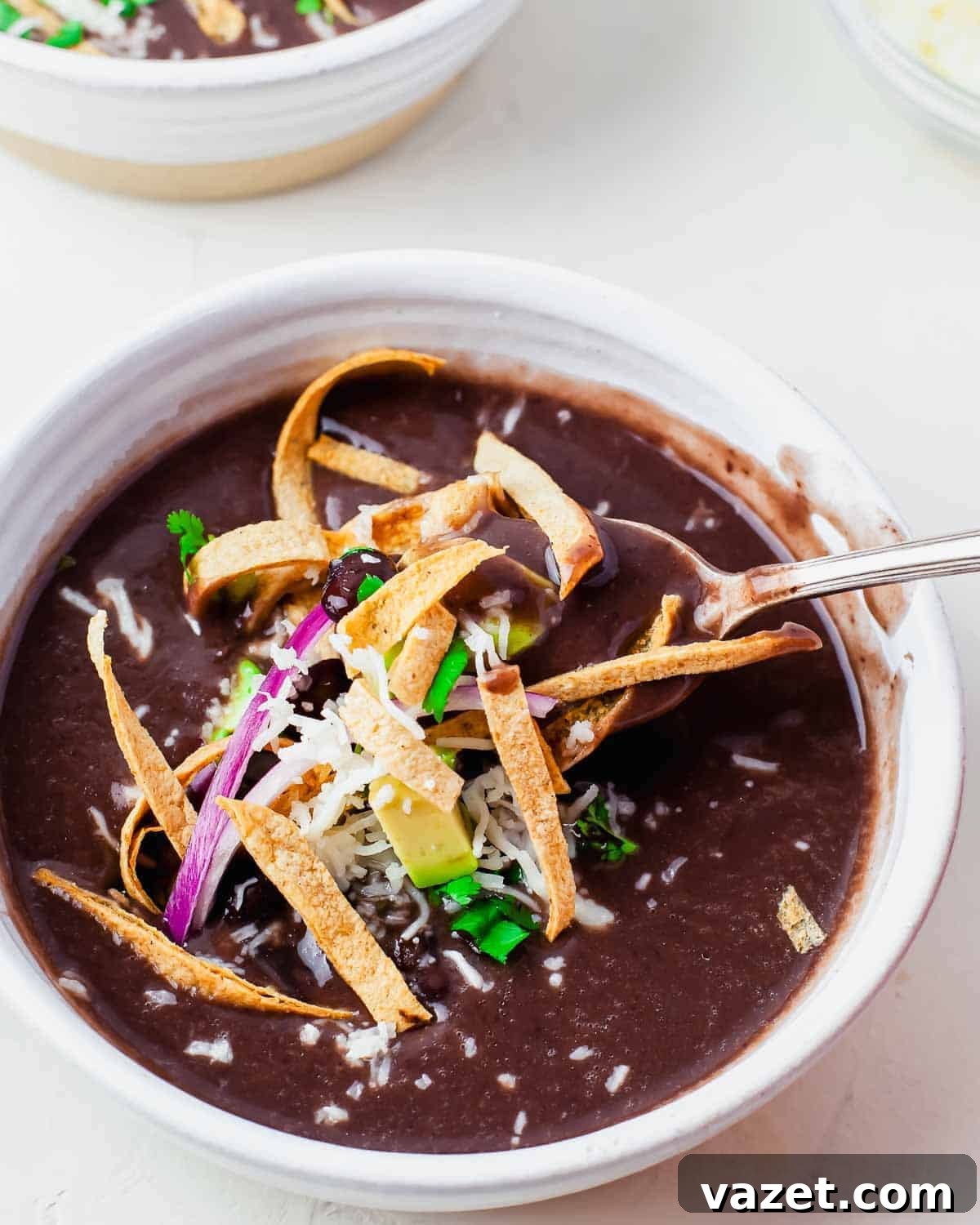 A serving of creamy Instant Pot black bean soup, garnished with golden-crisp tortilla strips, a sprinkle of shredded cheese, thinly sliced red onion, vibrant cilantro, and creamy avocado. A spoon gracefully lifts a mouthful of the flavorful soup.