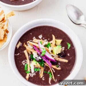 A bowl of Instant Pot Mexican black bean soup topped with tortilla strips, avocado, red onion, black beans, cheese, and chopped cilantro, with a spoon and extra tortilla strips on the side.