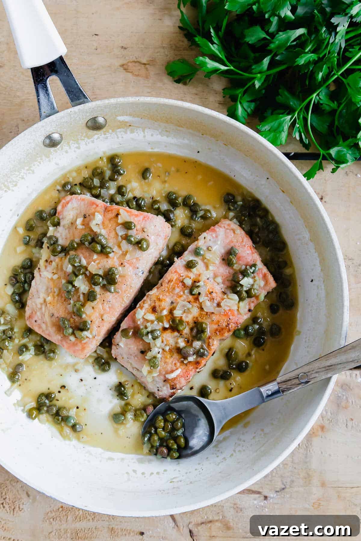 Lemon Butter Salmon Piccata with Capers and Parsley 7 Salmon piccata filets in a white skillet with a spoon.