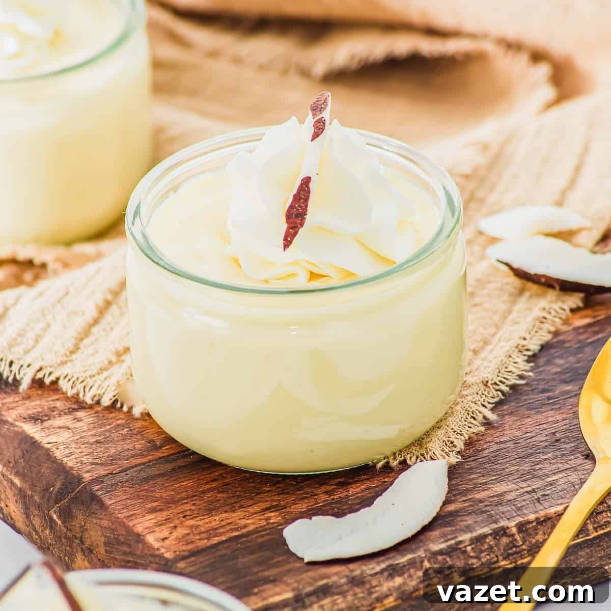 Creamy Coconut Pudding Recipe with Tropical Flavor 2 A small glass jar filled with coconut pudding, topped with whipped cream and a coconut shard, sits on a wooden board next to coconut flakes and a gold spoon.