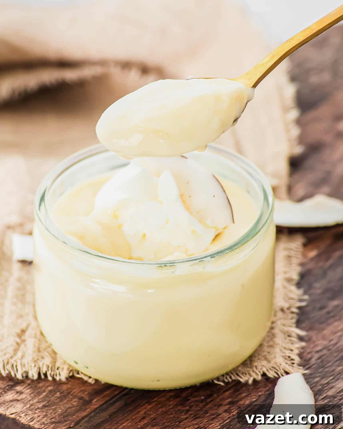 Creamy Coconut Pudding Recipe with Tropical Flavor 11 A spoon holding creamy, pale yellow coconut pudding hovers above an open glass jar filled with more custard, set on a piece of burlap atop a wooden surface. Coconut flakes are scattered nearby.