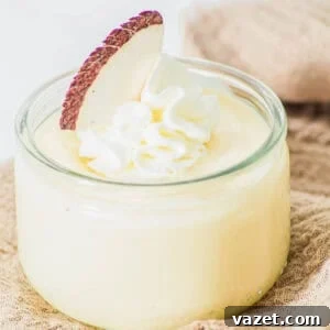Creamy Coconut Pudding Recipe with Tropical Flavor 13 A small glass jar filled with creamy yellow coconut pudding, topped with a swirl of whipped cream and a slice of fresh coconut, placed on a beige textured cloth.
