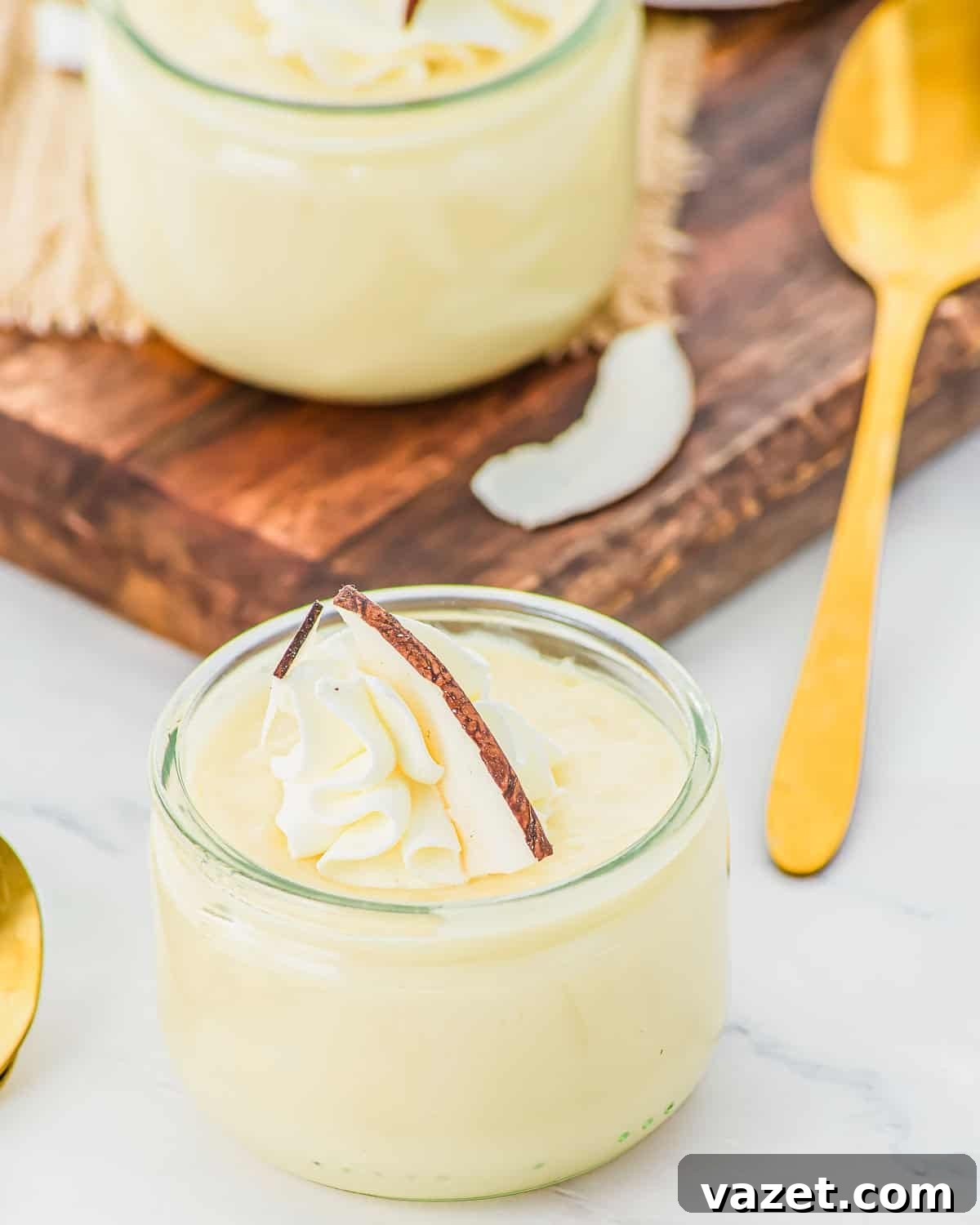 Creamy Coconut Pudding Recipe with Tropical Flavor 3 A small glass jar filled with creamy coconut pudding, topped with whipped cream and a slice of coconut, sits next to a gold spoon on a light surface. Another jar and a wooden board are in the background.