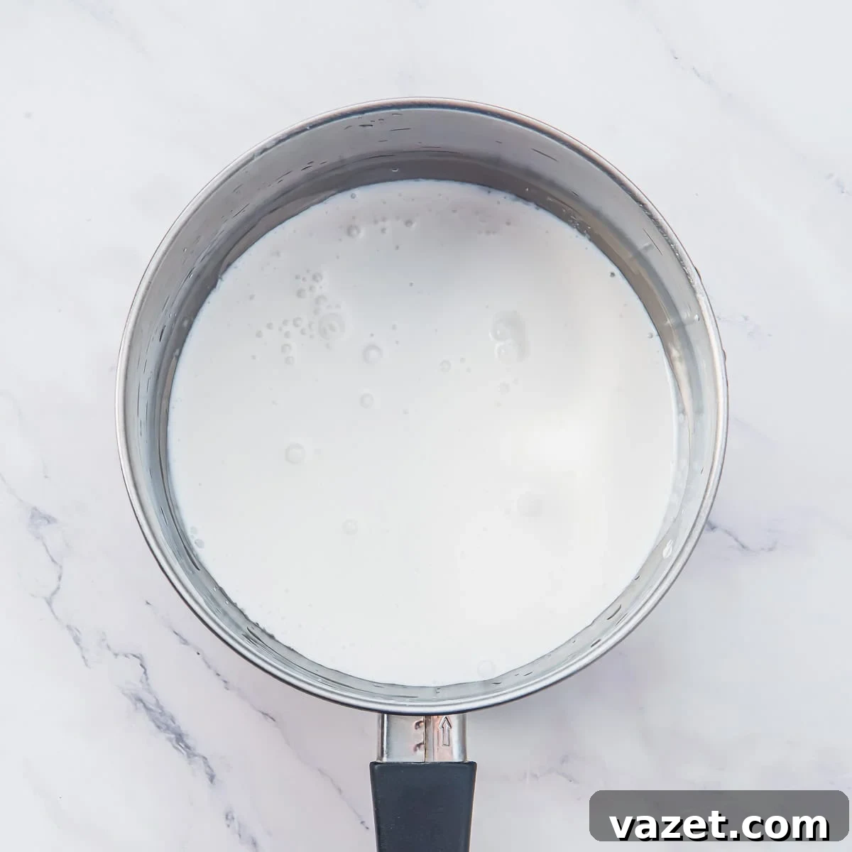 Creamy Coconut Pudding Recipe with Tropical Flavor 4 A stainless steel saucepan with a black handle, filled with coconut milk sits on a white marble surface.