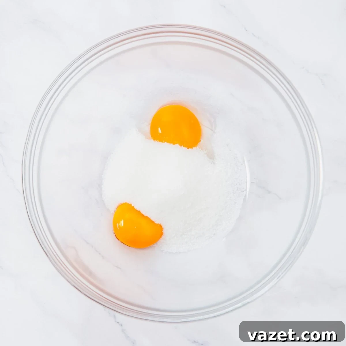 Creamy Coconut Pudding Recipe with Tropical Flavor 5 A glass bowl containing two egg yolks and a mound of granulated sugar, placed on a white marble surface.