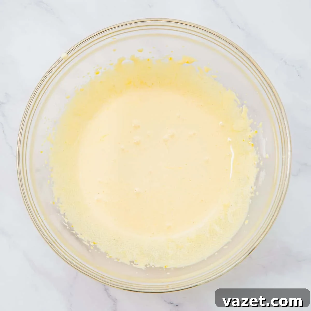 Creamy Coconut Pudding Recipe with Tropical Flavor 6 A glass bowl filled with whisked sugar and egg yolk mixture sits on a white marble surface. Small bubbles and flecks are visible around the edges of the mixture.