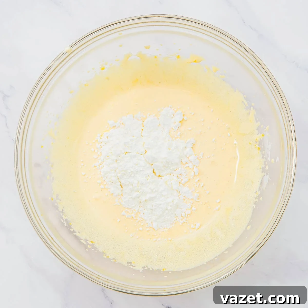 Creamy Coconut Pudding Recipe with Tropical Flavor 7 A glass bowl with whisked egg yolks and sugar with a pile of cornstarch in the center, on a white marble surface.