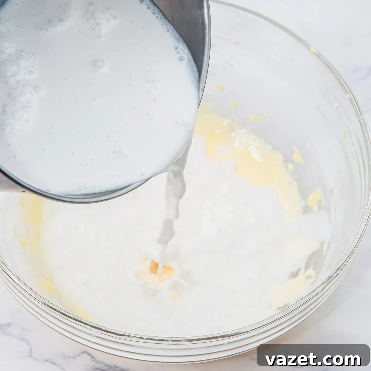 Creamy Coconut Pudding Recipe with Tropical Flavor 8 A metal bowl pours coconut milk into a glass mixing bowl containing a light yellow mixture, on a white marble surface.