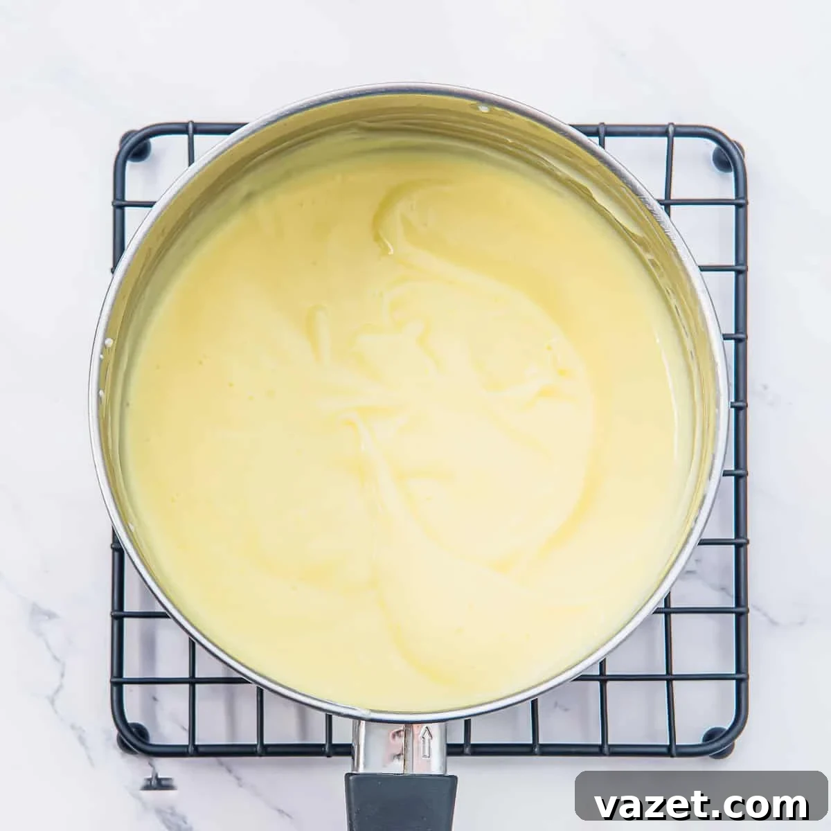 Creamy Coconut Pudding Recipe with Tropical Flavor 9 A saucepan filled with smooth, pale yellow pudding sits on a black wire cooling rack over a white marble surface.