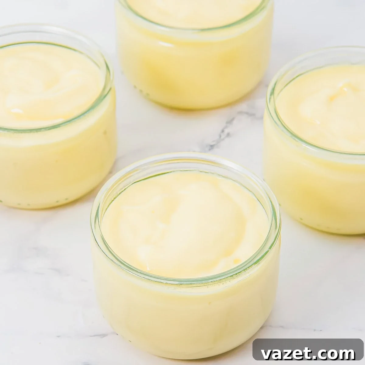 Creamy Coconut Pudding Recipe with Tropical Flavor 10 Four glass jars filled with creamy yellow coconut pudding are arranged on a white marble surface. The pudding has a smooth, thick texture and each jar is filled nearly to the top.