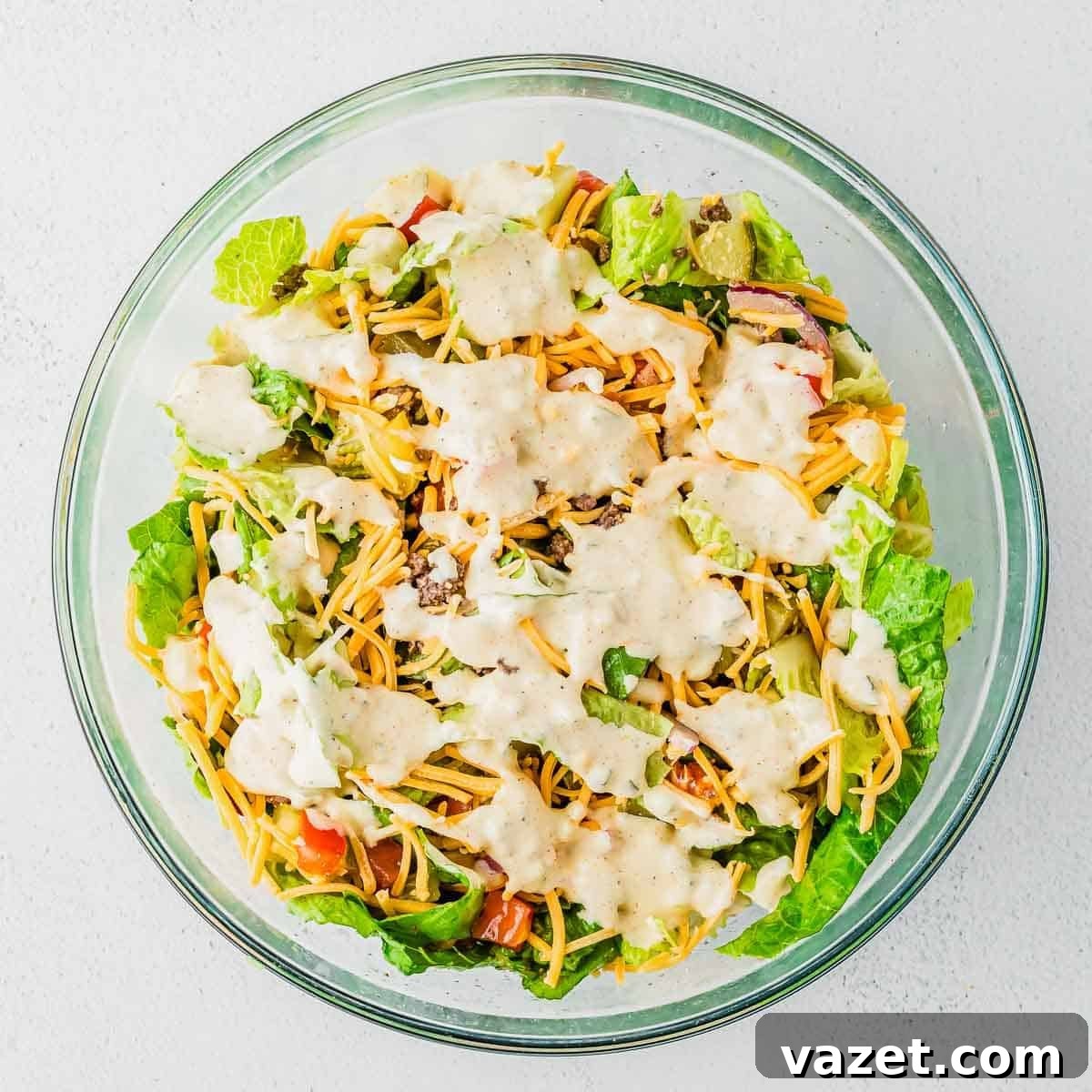 Big Mac Salad Recipe: Classic Cheeseburger Salad with Secret Sauce 11 A clear glass bowl filled with chopped lettuce, shredded cheddar cheese, tomatoes, and creamy dressing, on a white surface.