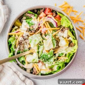 Big Mac Salad Recipe: Classic Cheeseburger Salad with Secret Sauce 13 A bowl of salad filled with lettuce, ground beef, shredded cheddar cheese, sliced red onions, diced tomatoes, pickles, and creamy dressing, with a fork in the bowl and some cheese scattered on the table.