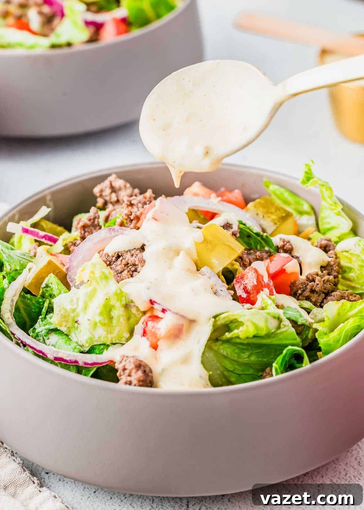 Big Mac Salad Recipe: Classic Cheeseburger Salad with Secret Sauce 4 A gray bowl filled with a salad containing ground beef, lettuce, tomato, pickles, and red onions, with creamy dressing being drizzled on top from a spoon above.