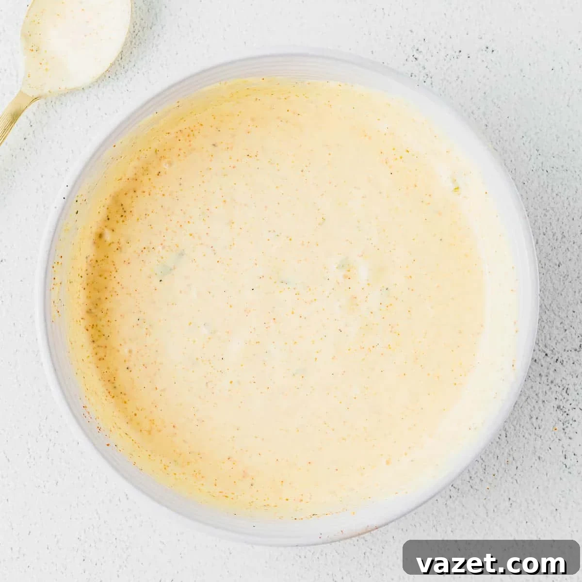 Big Mac Salad Recipe: Classic Cheeseburger Salad with Secret Sauce 6 A white bowl filled with a creamy, light burger sauce with specks of seasoning, placed on a light textured surface. A gold spoon with some sauce remains visible in the upper left corner.