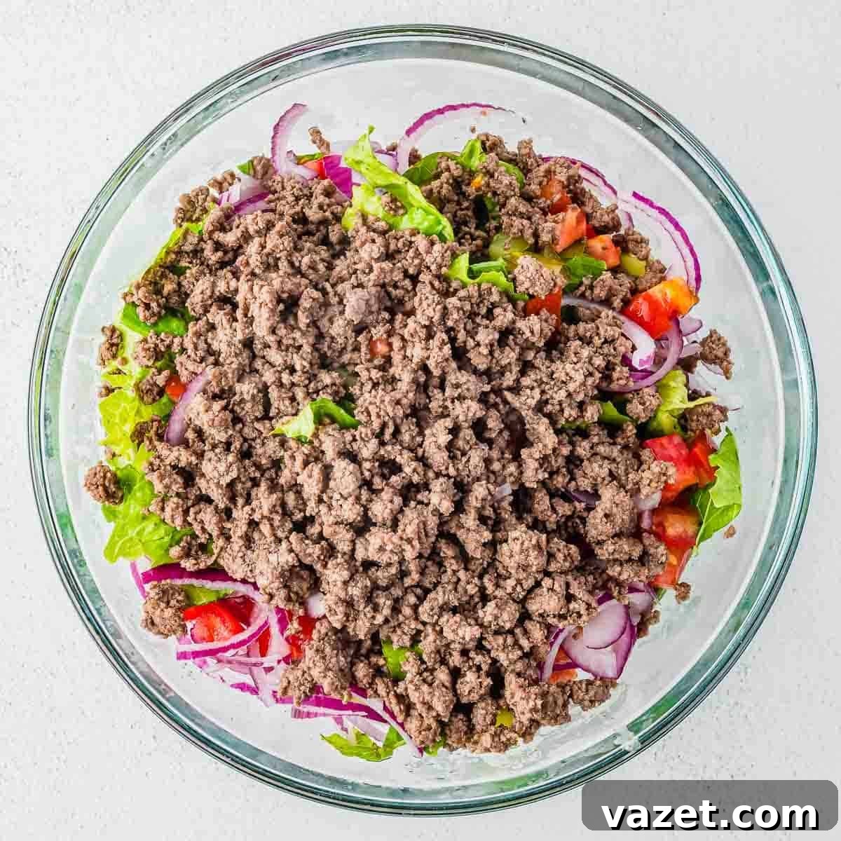 Big Mac Salad Recipe: Classic Cheeseburger Salad with Secret Sauce 9 A glass bowl filled with cooked ground beef on top of a salad with lettuce, sliced red onions, and diced tomatoes, all placed on a light-colored surface.