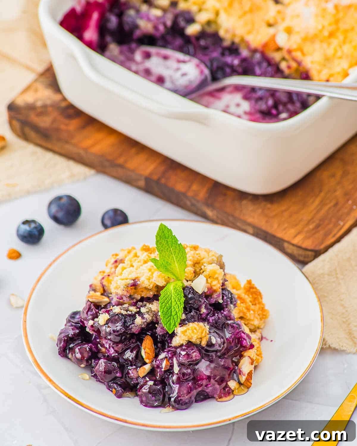 Classic Blueberry Cobbler Recipe with Cinnamon Crumble 2 A serving of blueberry cobbler topped with a mint sprig sits on a white plate, with a baking dish of the dessert and a spoon in the background. Blueberries and chopped nuts are scattered around.