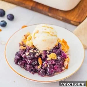 Classic Blueberry Cobbler Recipe with Cinnamon Crumble 12 A white plate with a serving of blueberry cobbler topped with a scoop of vanilla ice cream, garnished with sliced almonds. Fresh blueberries and a wooden board are in the background.