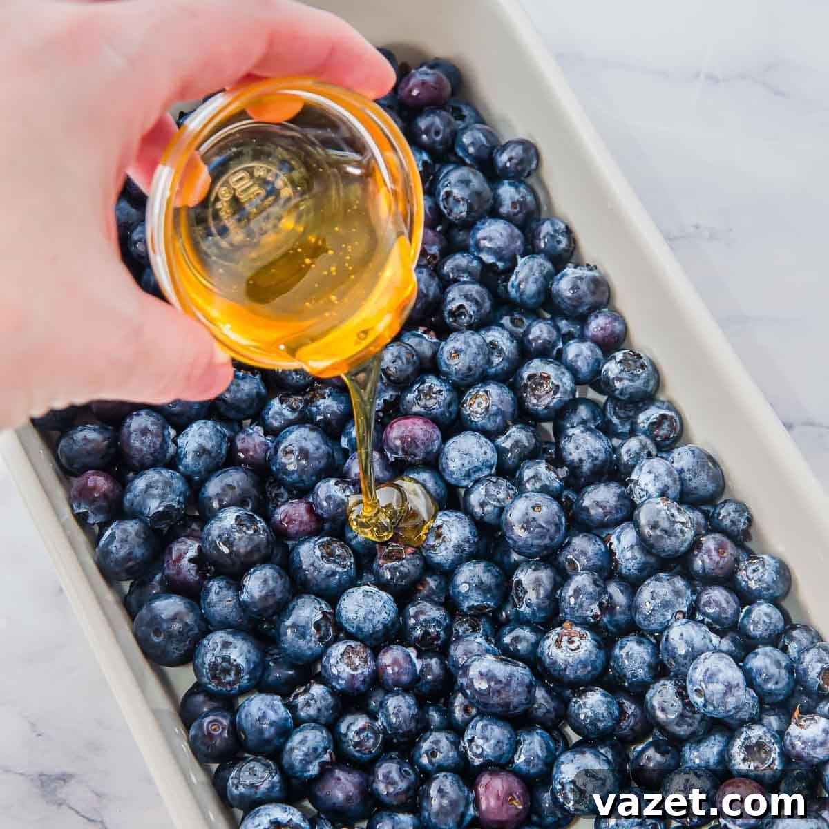 Classic Blueberry Cobbler Recipe with Cinnamon Crumble 3 A hand pours golden honey from a small glass bowl over fresh blueberries arranged in a rectangular white baking dish.