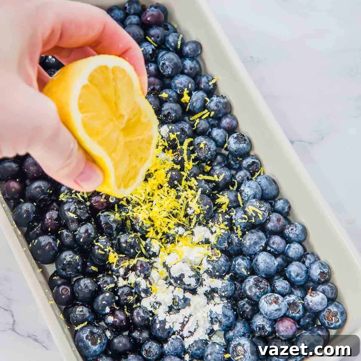 Classic Blueberry Cobbler Recipe with Cinnamon Crumble 4 A hand squeezes a lemon half over a baking dish filled with fresh blueberries, lemon zest, and a sprinkle of flour.